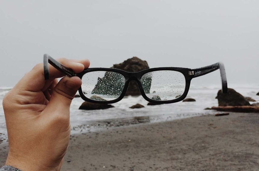 Hand holding rain-speckled glasses at rocky beach